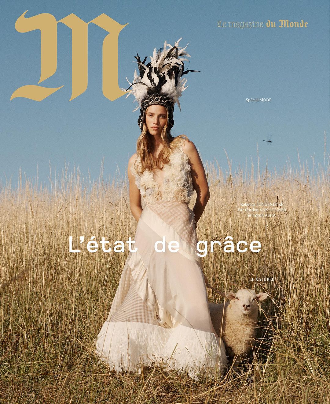 Magazine cover featuring a woman in couture gown with feather headdress standing in a field next to a sheep on a sunny day.