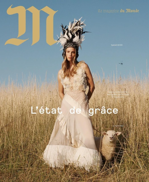 Magazine cover featuring a woman in couture gown with feather headdress standing in a field next to a sheep on a sunny day.