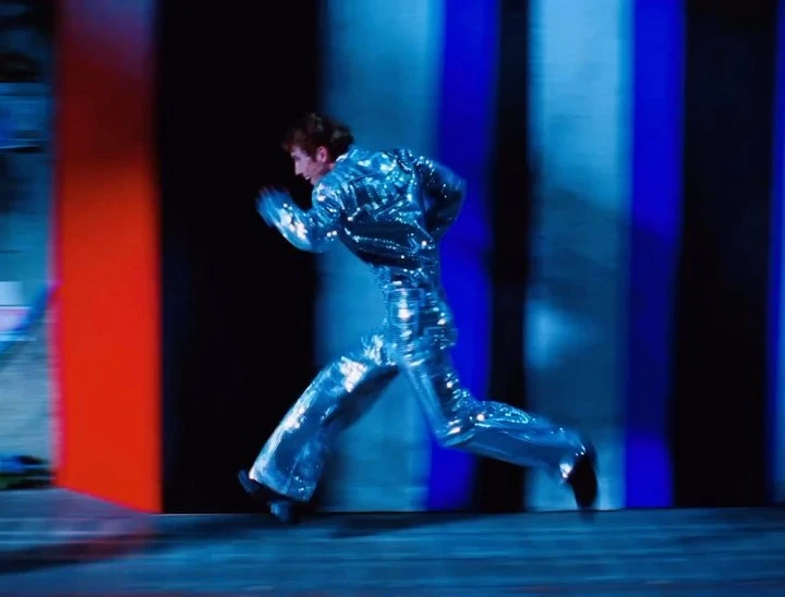 Person in a shiny silver outfit runs in a blurry indoor space with a blue, black, and red striped background, long exposure.