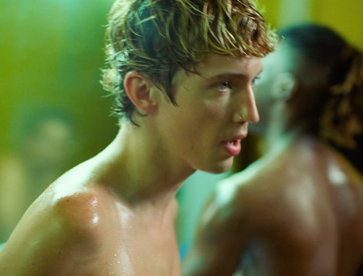 Close-up on an adult male with blonde hair and a nose ring, sweat glistening on his skin, against a muted green and yellow background.