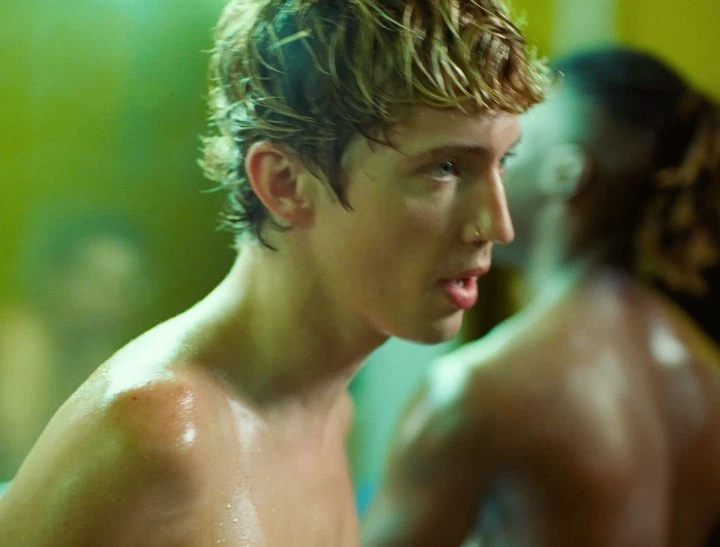 Close-up on an adult male with blonde hair and a nose ring, sweat glistening on his skin, against a muted green and yellow background.