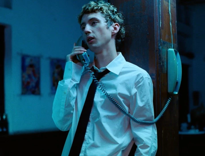 Portrait of a young adult male on a vintage telephone indoors, wearing an unbuttoned white shirt and black tie in dim blue lighting.