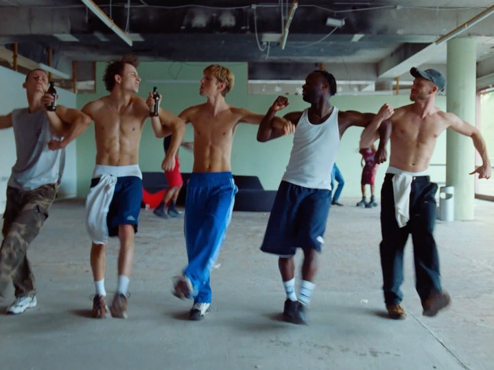 Five shirtless men dancing in a bright, open space with beer bottles in hand, capturing a candid moment of youthful energy.