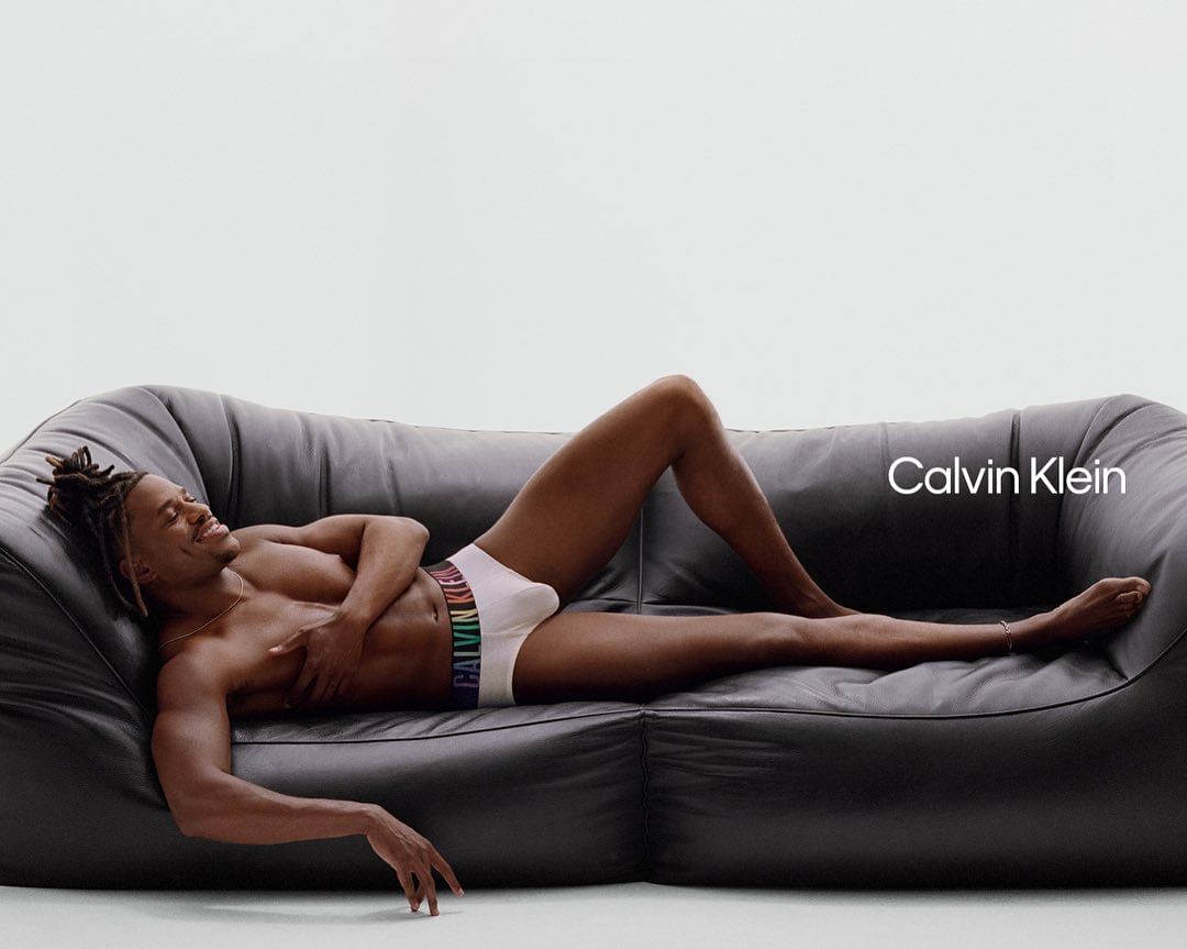An African-American male reclines on a black leather sofa in white Calvin Klein underwear with a multicolored waistband, set against a white background.