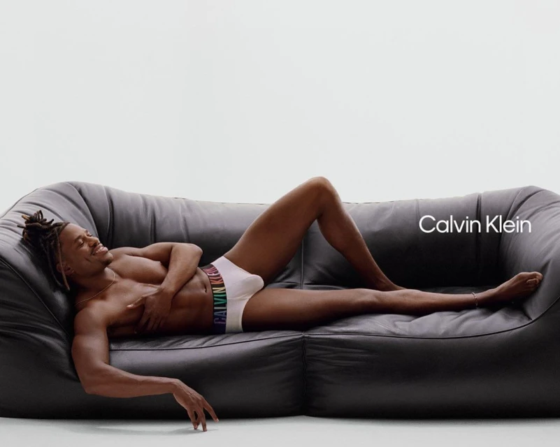 An African-American male reclines on a black leather sofa in white Calvin Klein underwear with a multicolored waistband, set against a white background.