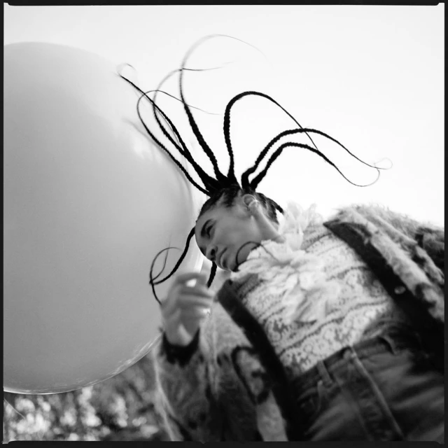 Artistic black and white portrait of a person with braided hair holding a large balloon, wearing patterned clothing, outdoors.