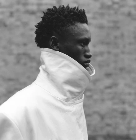 Profile portrait of a black man wearing a high-collared white jacket against a textured brick wall in Paris.