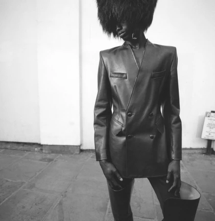 Black and white fashion portrait of an African American model in a leather suit and large furry hat standing indoors against a white wall.