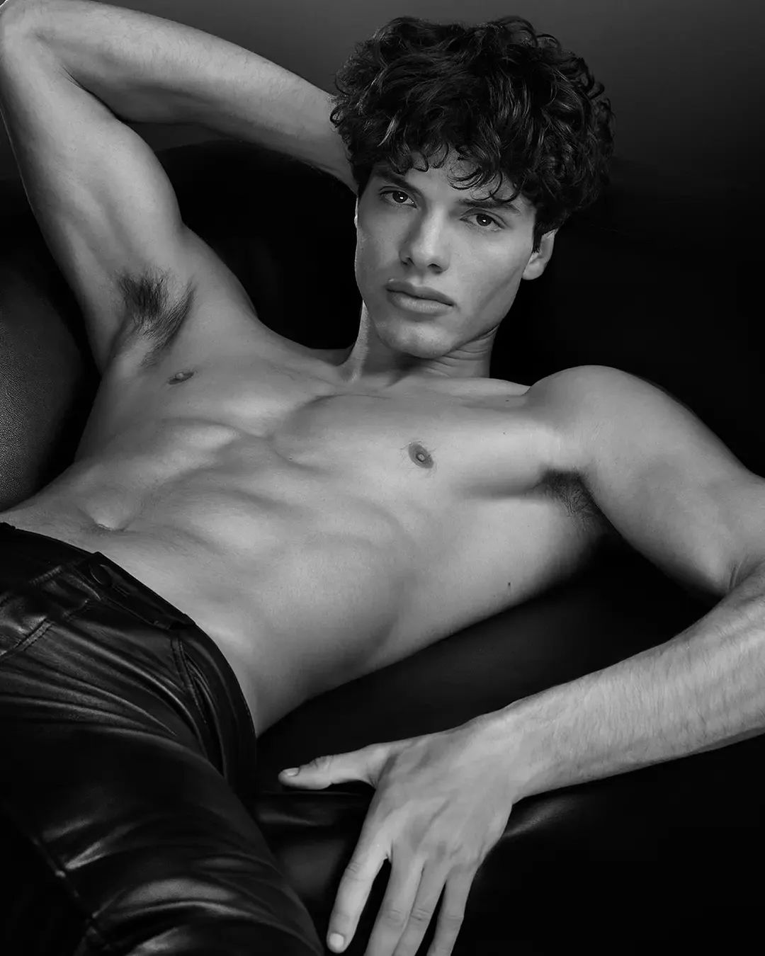 Black and white portrait of a muscular adult male lying on black leather. Features include curly dark hair, leather pants, and studio lighting.