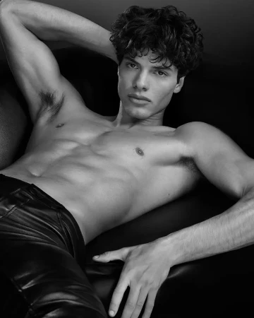 Black and white portrait of a muscular adult male lying on black leather. Features include curly dark hair, leather pants, and studio lighting.