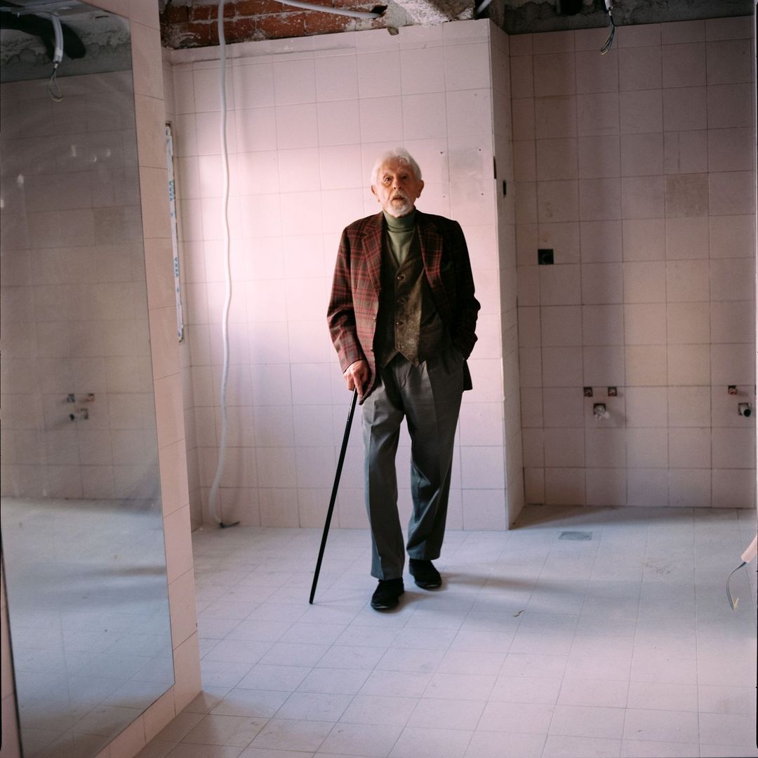 Elderly man with cane walks in tiled room with exposed ceiling. He wears a tweed jacket, vest, grey pants, and black shoes.