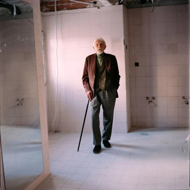 Elderly man with cane walks in tiled room with exposed ceiling. He wears a tweed jacket, vest, grey pants, and black shoes.