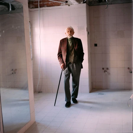 Elderly man with cane walks in tiled room with exposed ceiling. He wears a tweed jacket, vest, grey pants, and black shoes.