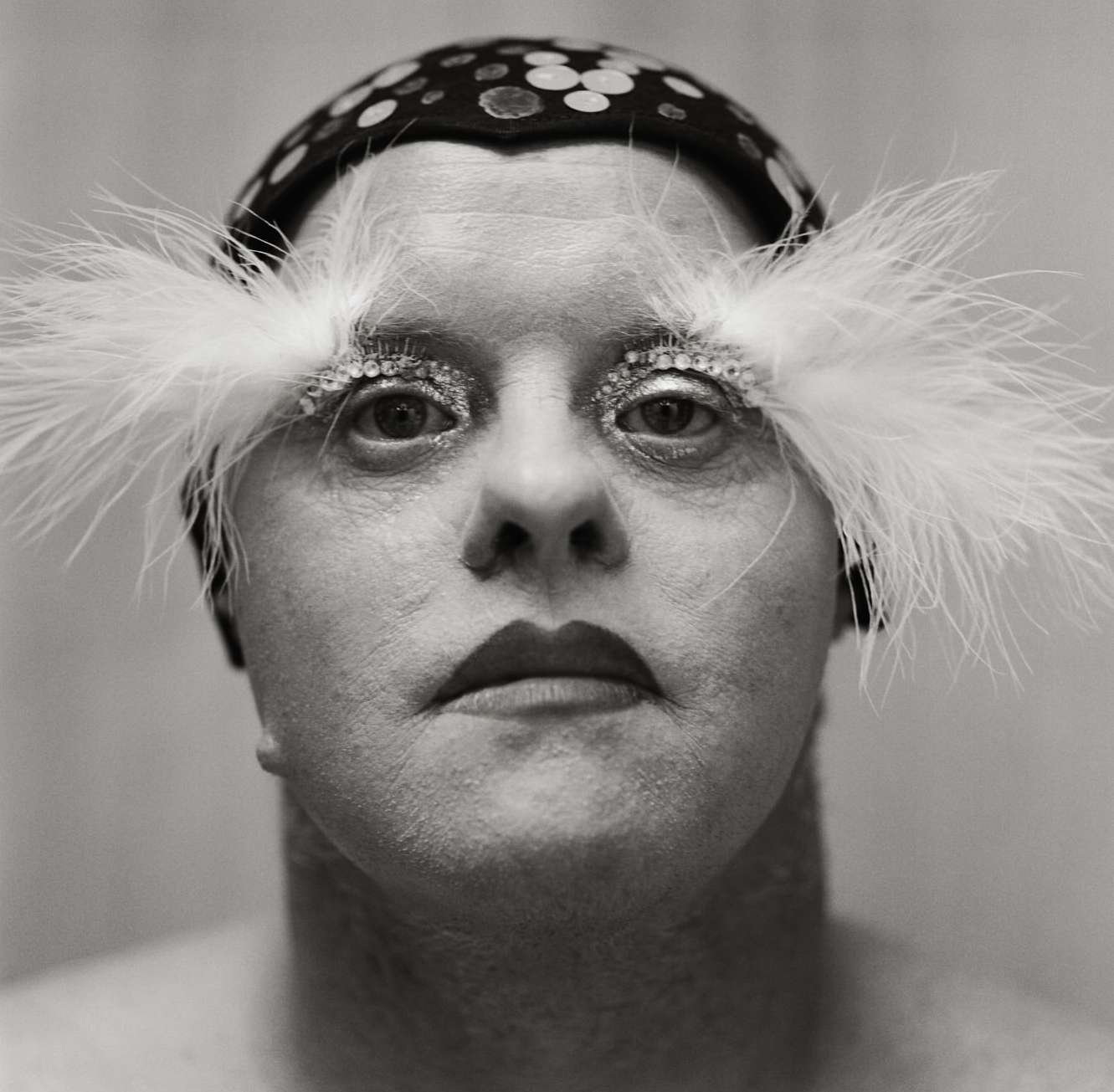 Close-up portrait of an adult with decorative eye makeup, a feathered headpiece, and a polka-dot hat against a neutral monochrome background.