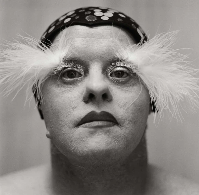 Close-up portrait of an adult with decorative eye makeup, a feathered headpiece, and a polka-dot hat against a neutral monochrome background.