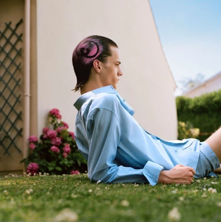 Young man in a light blue shirt reclining on grass with pink-dyed hair featuring a shaved black swirl design