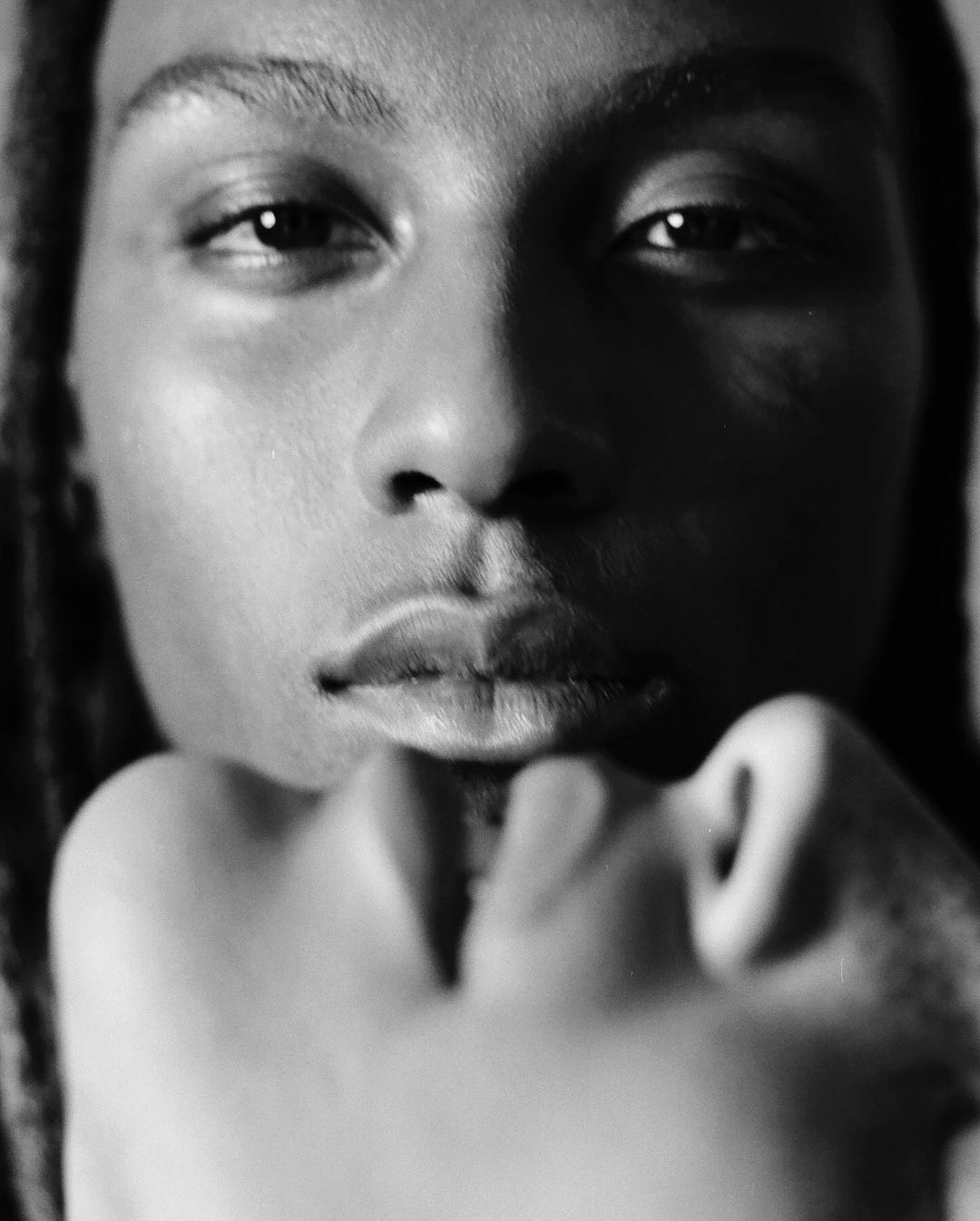 Monochrome portrait of a person with dreadlocks resting their chin on their hand. The image shows a close-up shot, eye-level view.