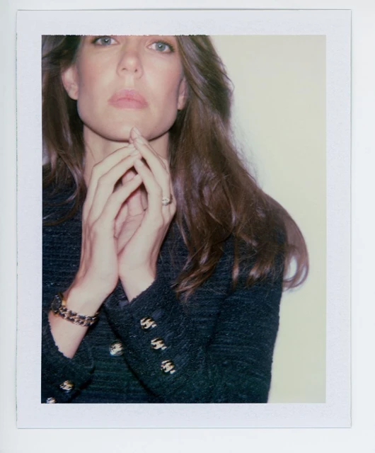 Portrait of an adult female in a black jacket with gold buttons, posing with her hands near her face in an indoor setting.