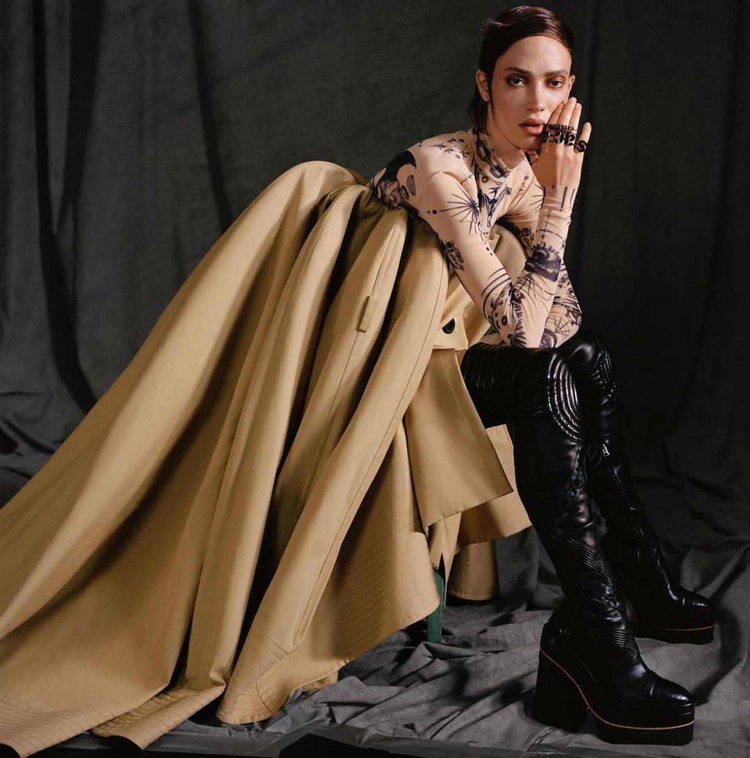 Woman poses in long-sleeved printed top, black leather boots, and tan trench coat in a studio setting with dark gray backdrop.