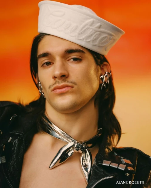 Close-up studio shot of a stylish adult male wearing a sailor hat, jewelry, a silver scarf, and a leather jacket against an orange background.
