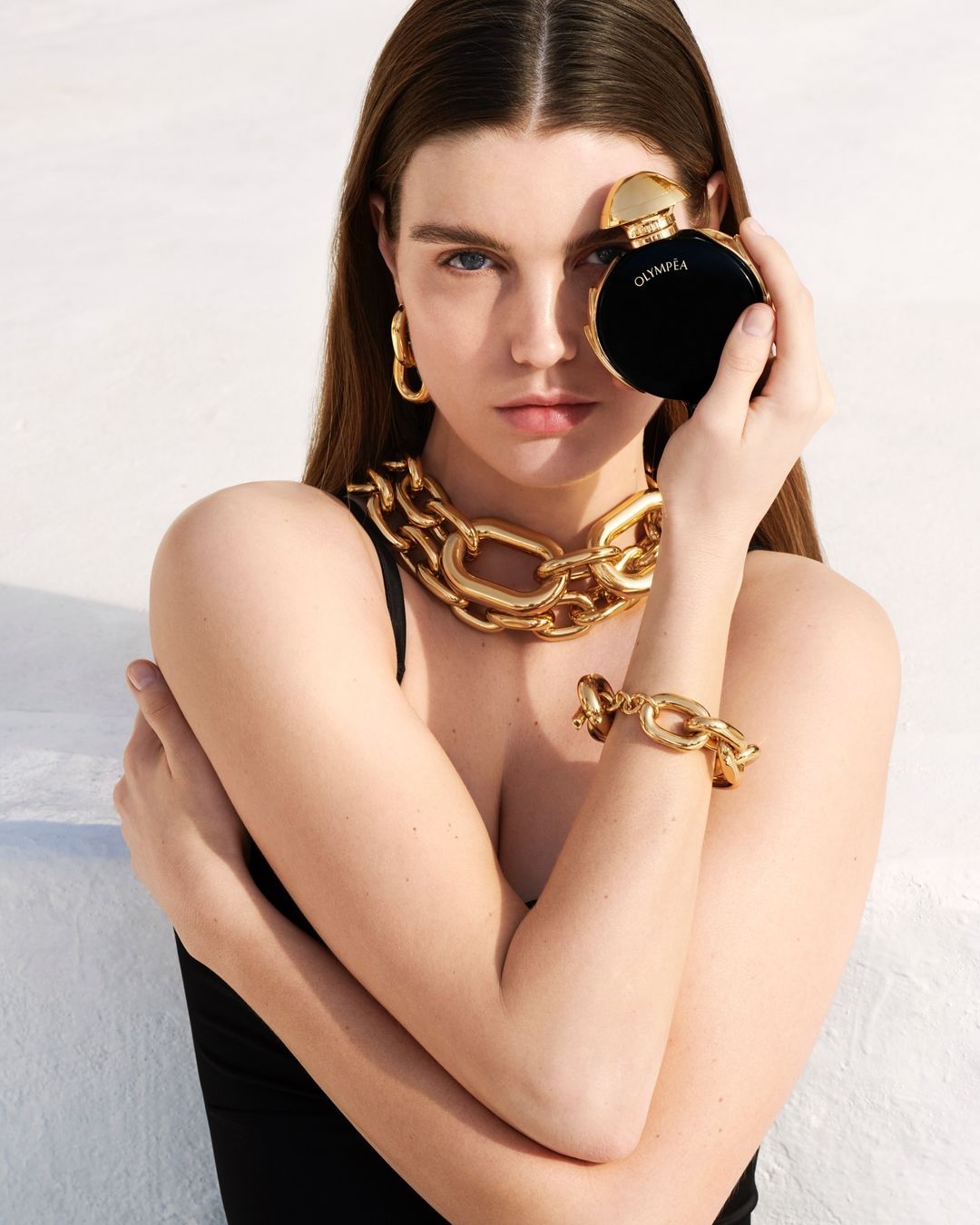 A young woman poses with a black perfume bottle, wearing a gold necklace and earrings against a bright white background in a close-up shot.