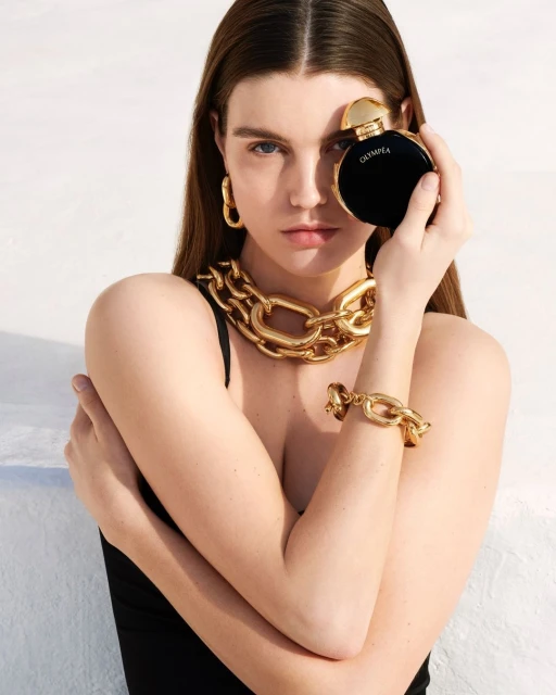 A young woman poses with a black perfume bottle, wearing a gold necklace and earrings against a bright white background in a close-up shot.