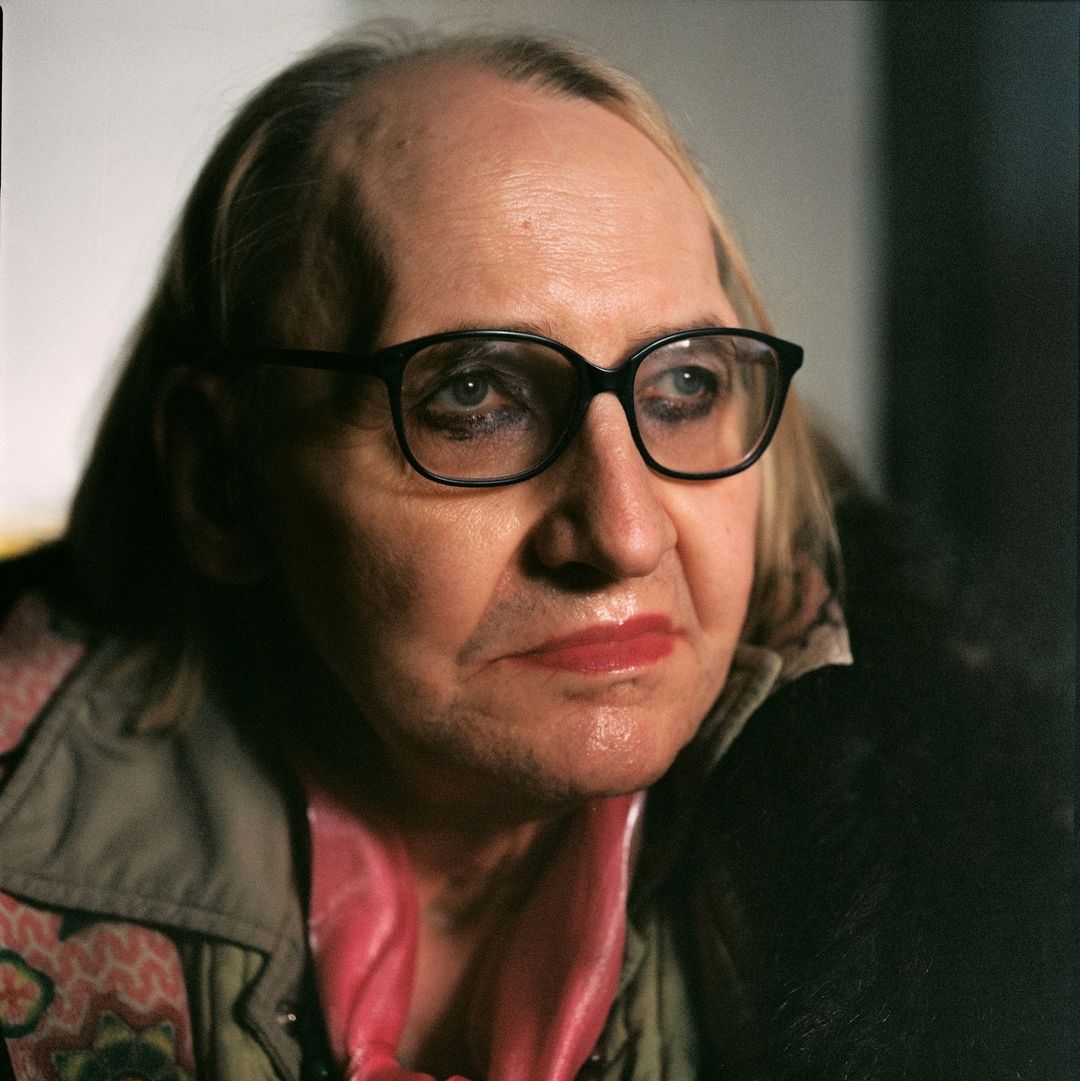 Close-up portrait of an elderly person wearing black glasses, pink scarf, and textured jacket by photographer Gorka Postigo.