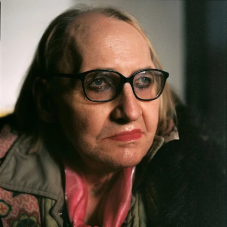 Close-up portrait of an elderly person wearing black glasses, pink scarf, and textured jacket by photographer Gorka Postigo.