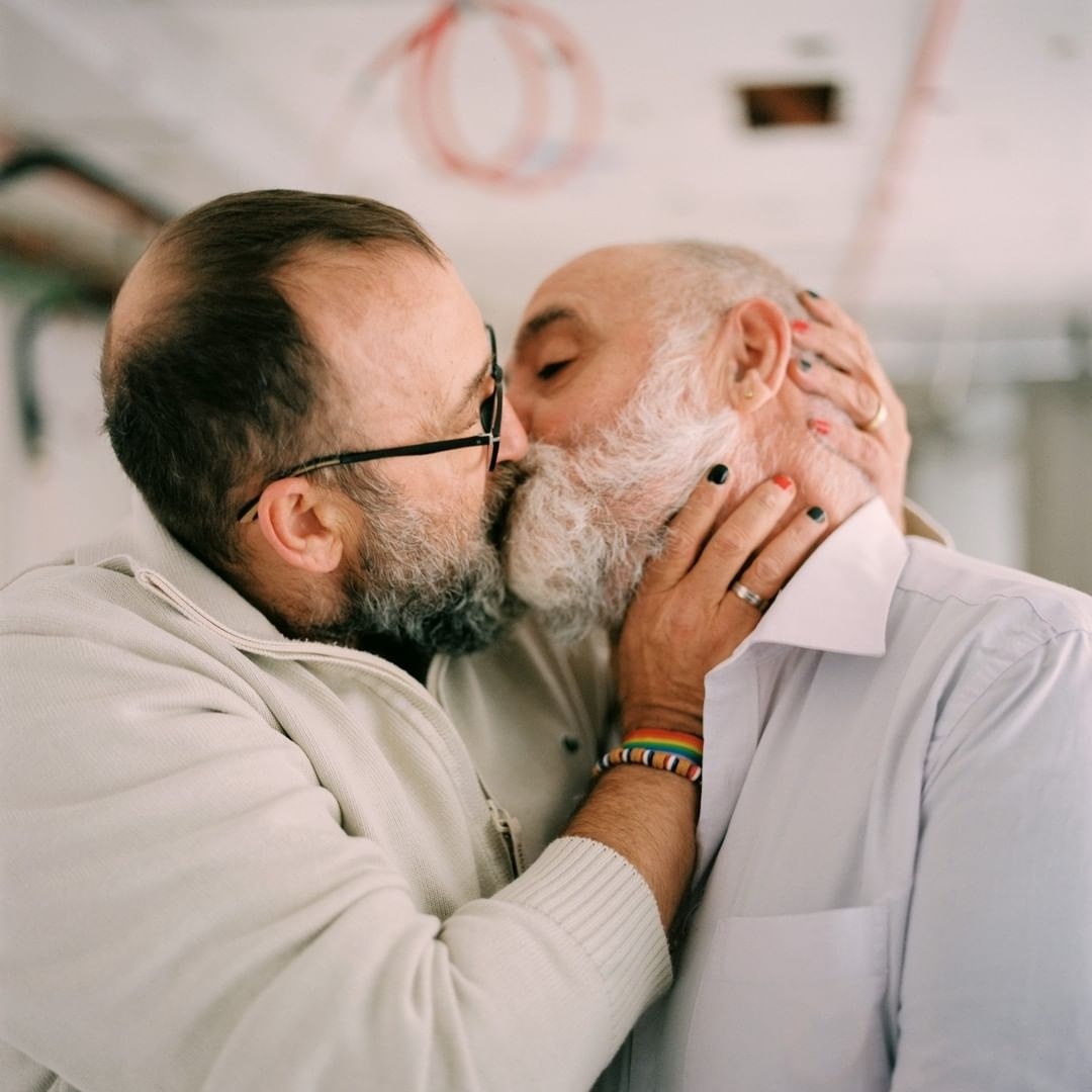 Close-up of two elderly men kissing, showcasing their beards, glasses, and casual attire in a simple indoor environment.