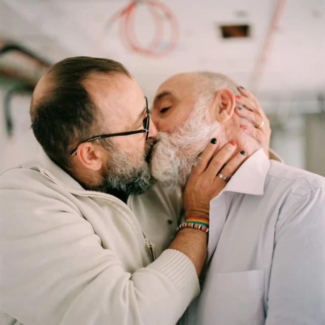 Close-up of two elderly men kissing, showcasing their beards, glasses, and casual attire in a simple indoor environment.