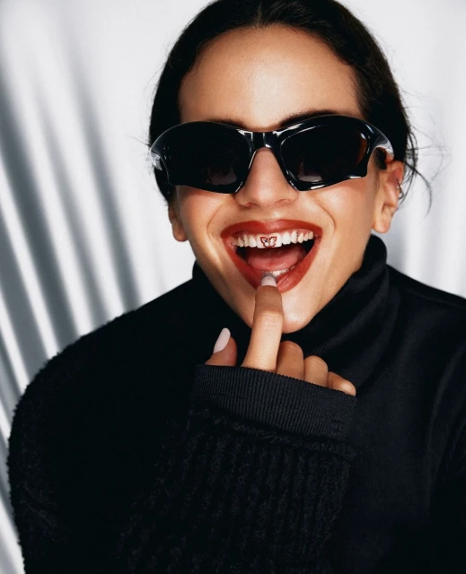 Close-up of a woman with sunglasses, red lipstick, and a tooth gem, wearing a black turtleneck sweater against a bright backdrop.