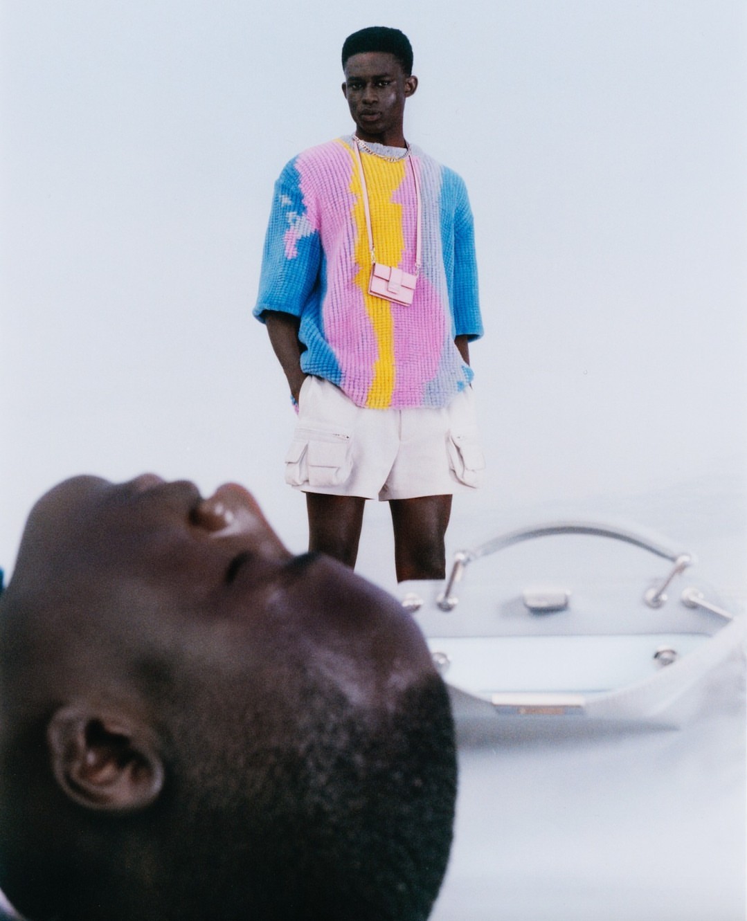 Fashion shot of a Black male model wearing colorful knit shirt and shorts with a small purse standing next to a silver object.