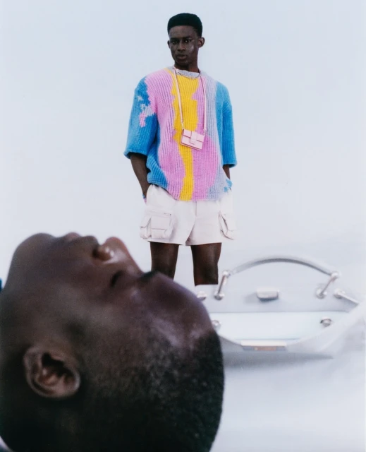 Fashion shot of a Black male model wearing colorful knit shirt and shorts with a small purse standing next to a silver object.
