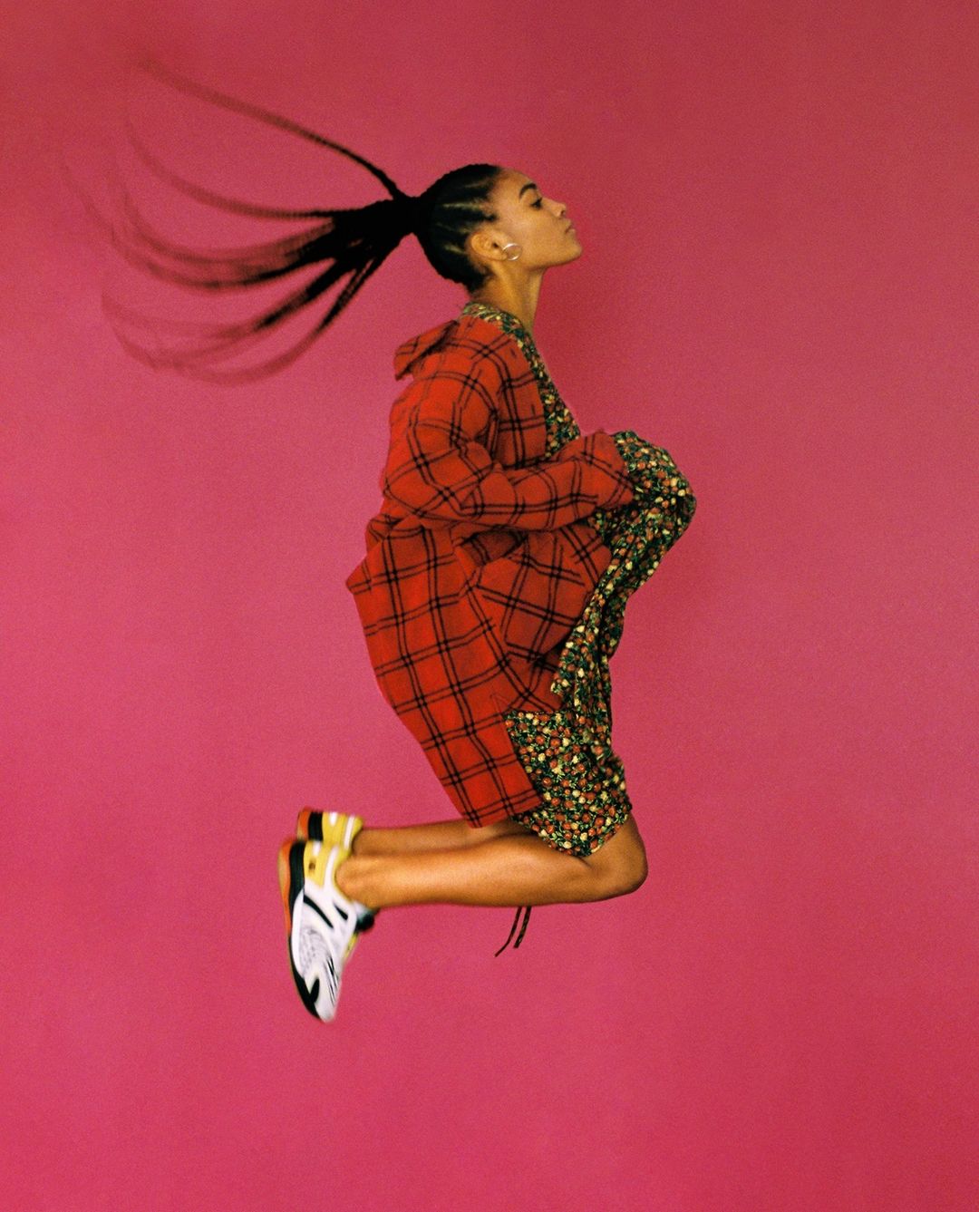 A profile shot of an adult woman jumping with a red plaid jacket and floral shorts against a solid pink backdrop in a vibrant studio.