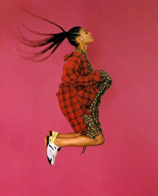 A profile shot of an adult woman jumping with a red plaid jacket and floral shorts against a solid pink backdrop in a vibrant studio.