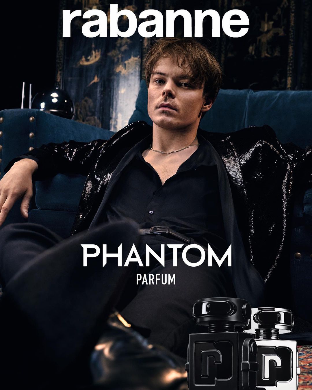 Fashion shot for Rabanne featuring a young adult male model in a black outfit sitting on a blue velvet sofa next to perfume bottles.
