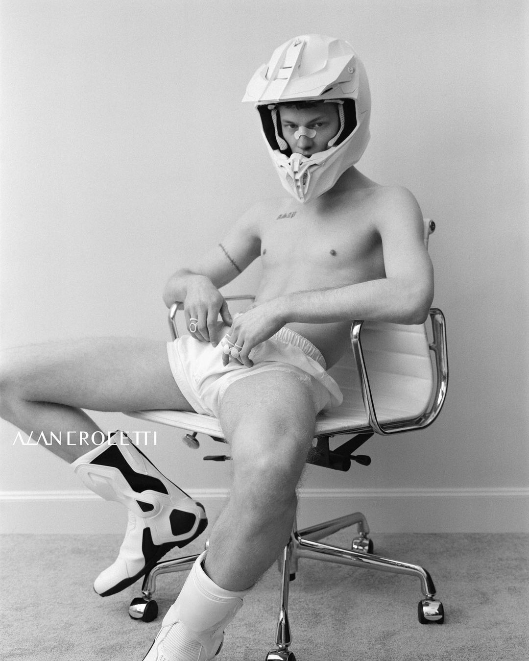Monochrome portrait of a man in a motorcycle helmet and boots, seated in an office chair, set against a plain background.
