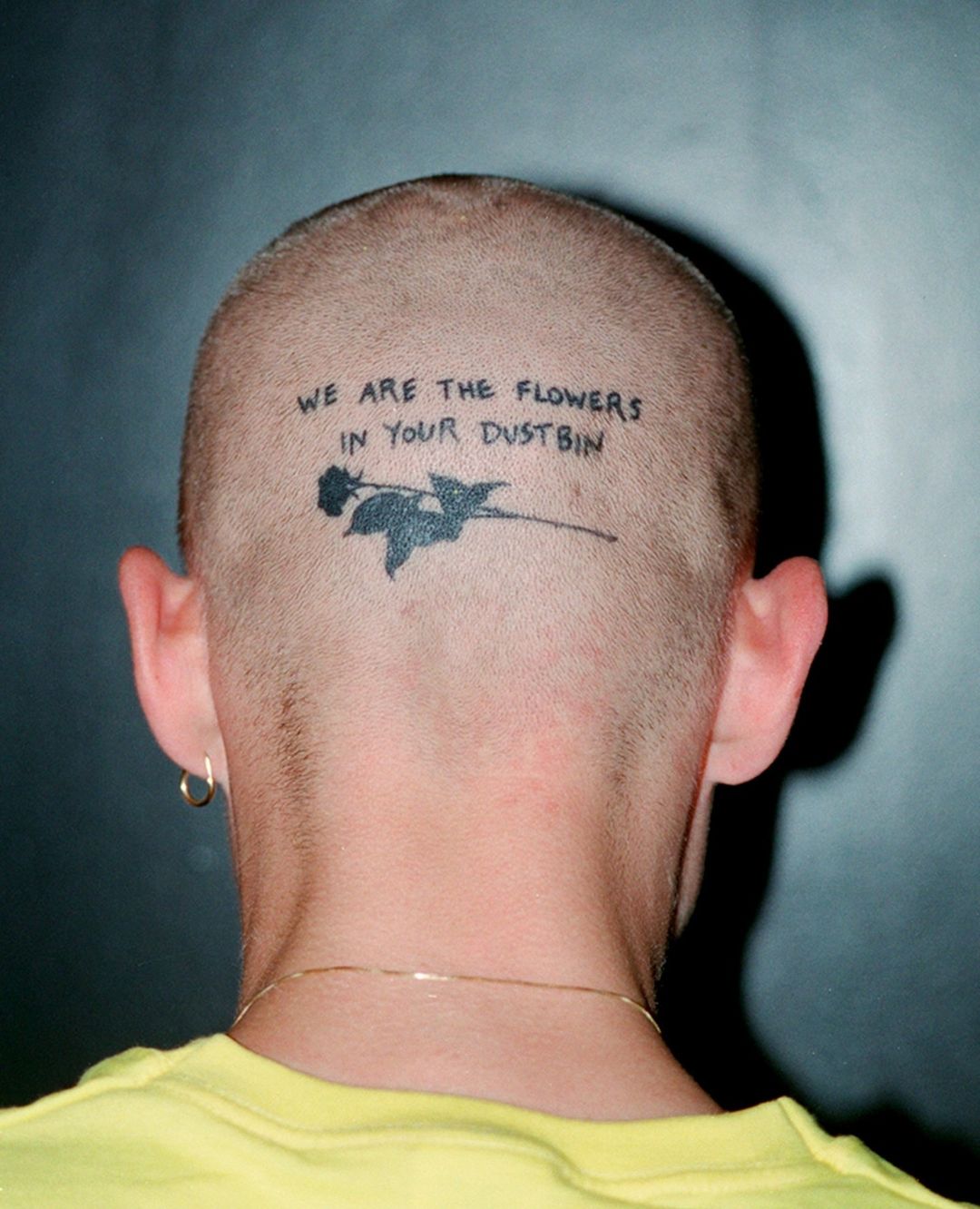 Adult male's shaved head with a black tattoo of a rose and the text 'We are the flowers in your dustbin' against a gradient gray background.