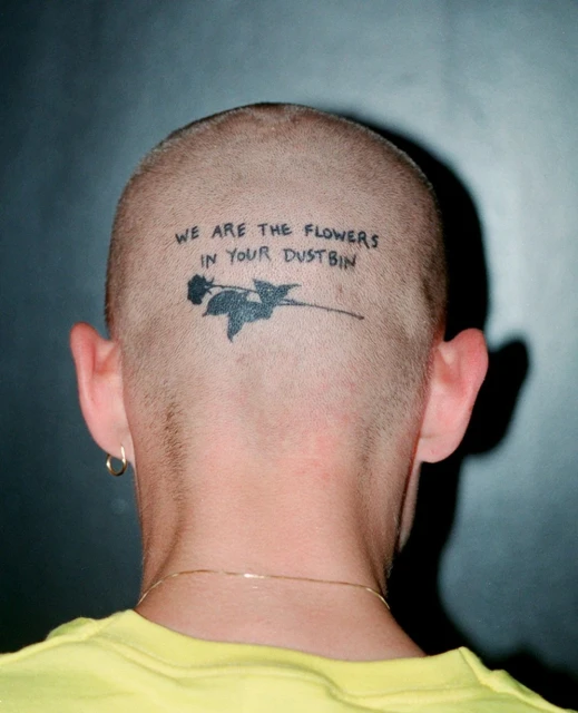 Adult male's shaved head with a black tattoo of a rose and the text 'We are the flowers in your dustbin' against a gradient gray background.