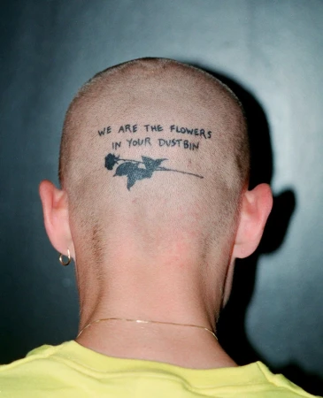Adult male's shaved head with a black tattoo of a rose and the text 'We are the flowers in your dustbin' against a gradient gray background.