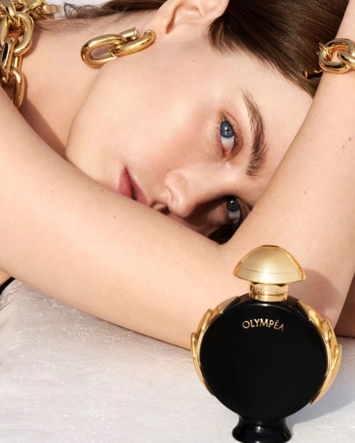 Close-up of a woman with fair skin and blue eyes wearing gold jewelry, positioned near a black and gold perfume bottle.