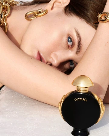 Close-up of a woman with fair skin and blue eyes wearing gold jewelry, positioned near a black and gold perfume bottle.