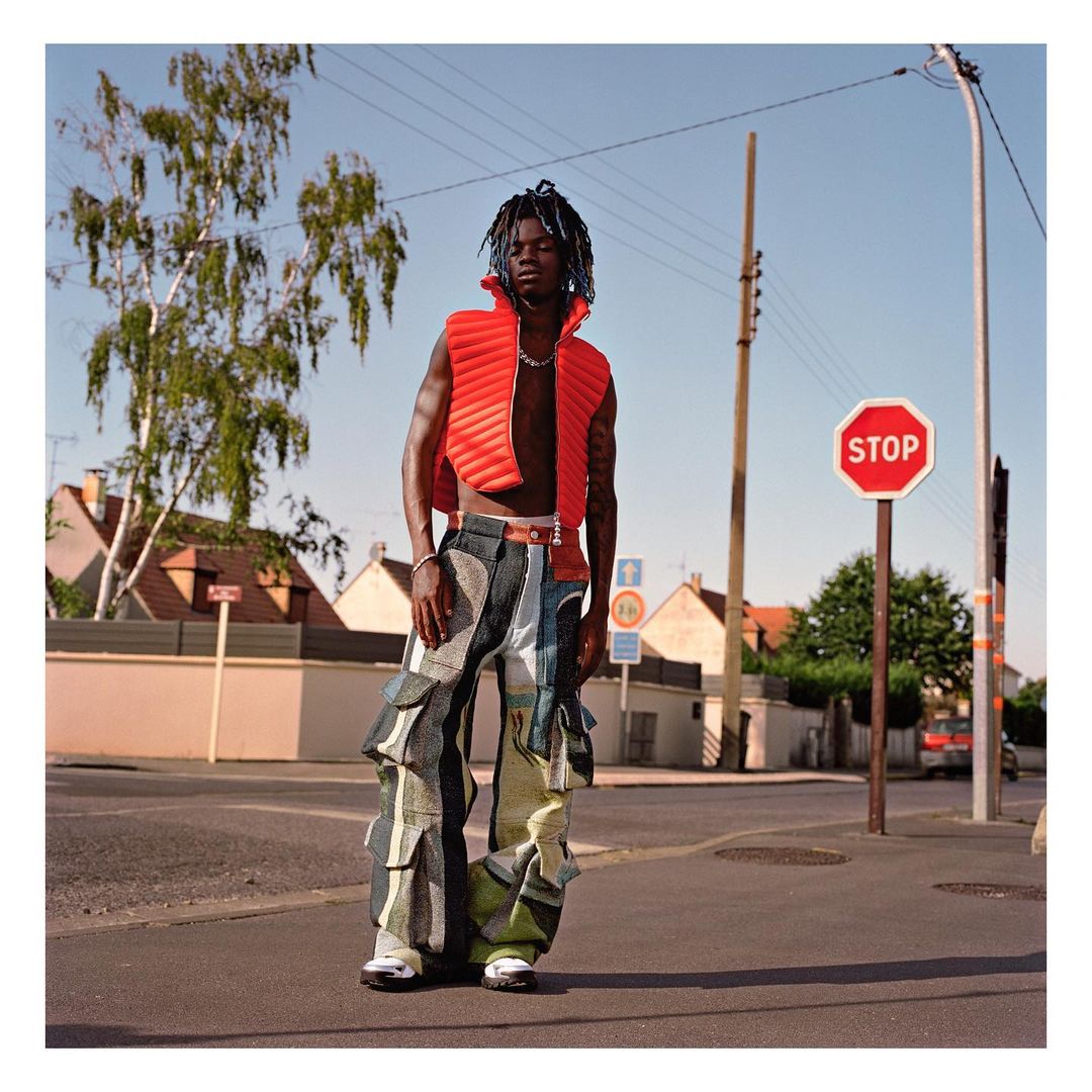 A full shot of a young adult male in a red vest and patterned pants standing on a street near a stop sign during the daytime.