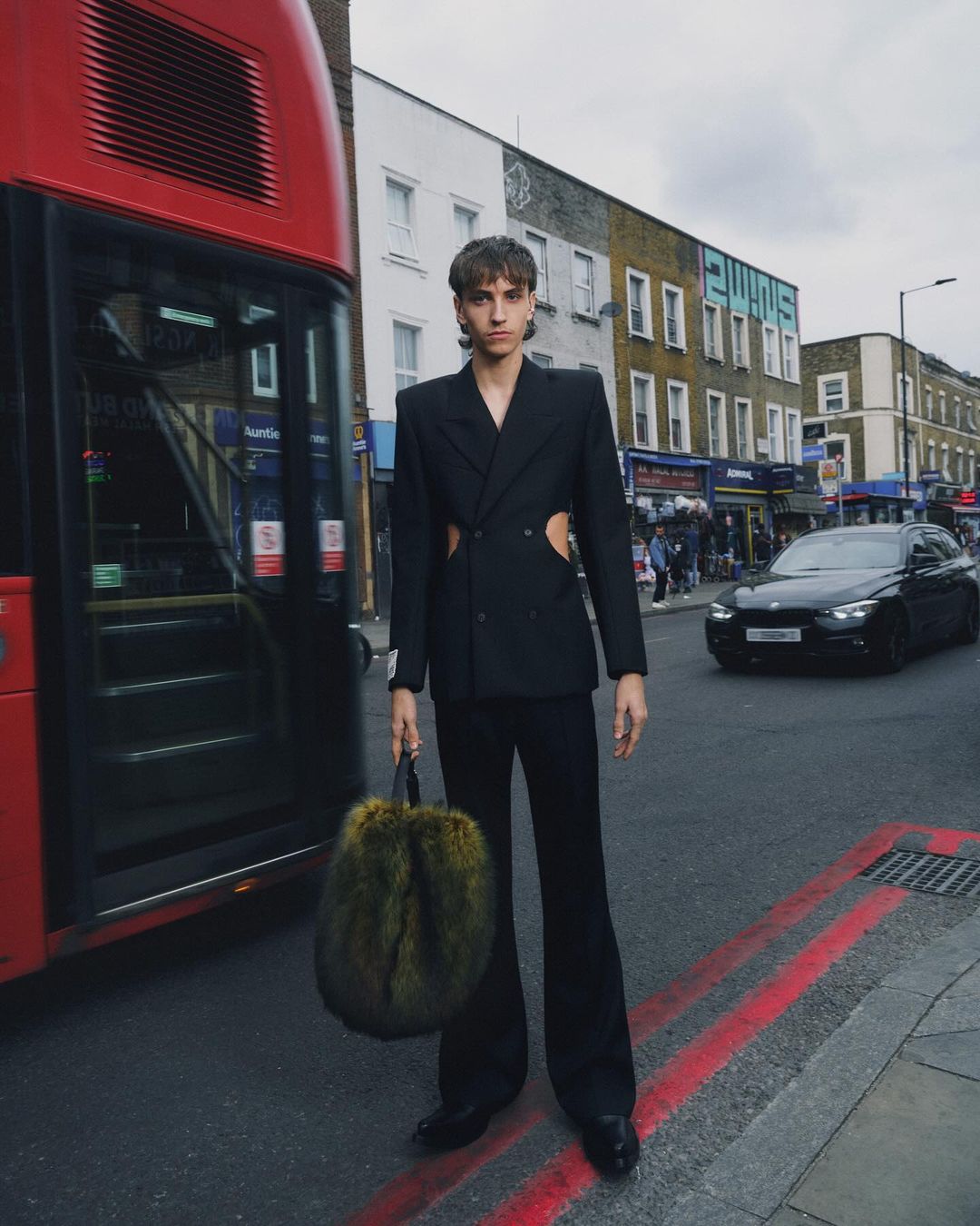 Fashion model in a black suit stands on a city street near a red double-decker bus, holding a fur handbag in an editorial style.