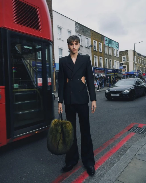 Fashion model in a black suit stands on a city street near a red double-decker bus, holding a fur handbag in an editorial style.