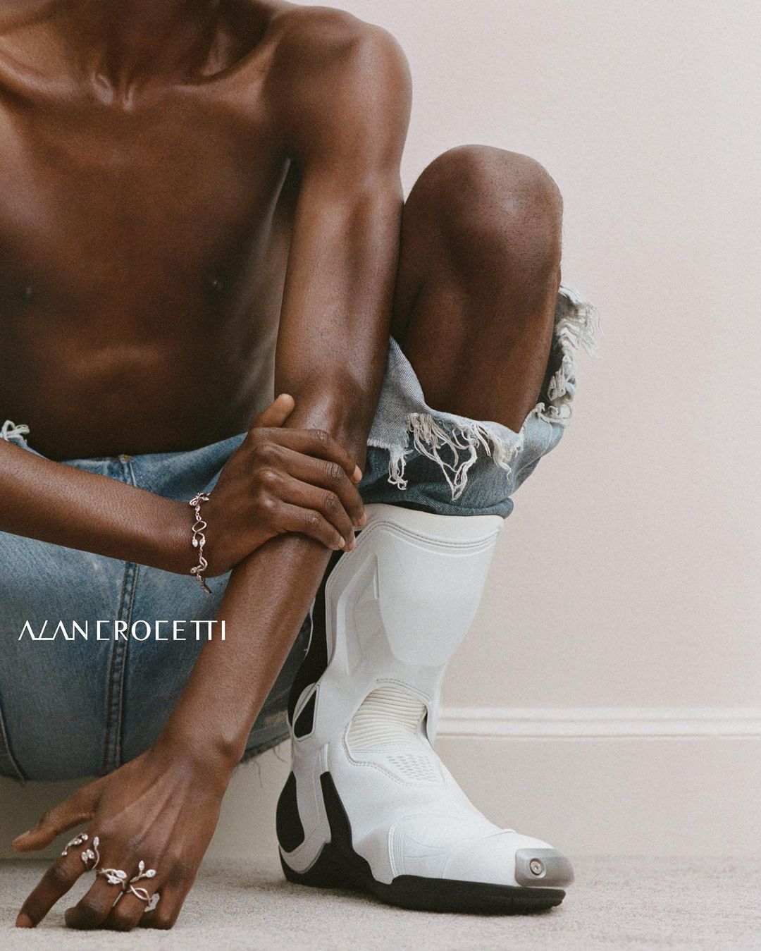 Dark-skinned shirtless person in ripped denim with white moto boot and jewelry. Fashion-focused portrait in a bright, neutral studio.