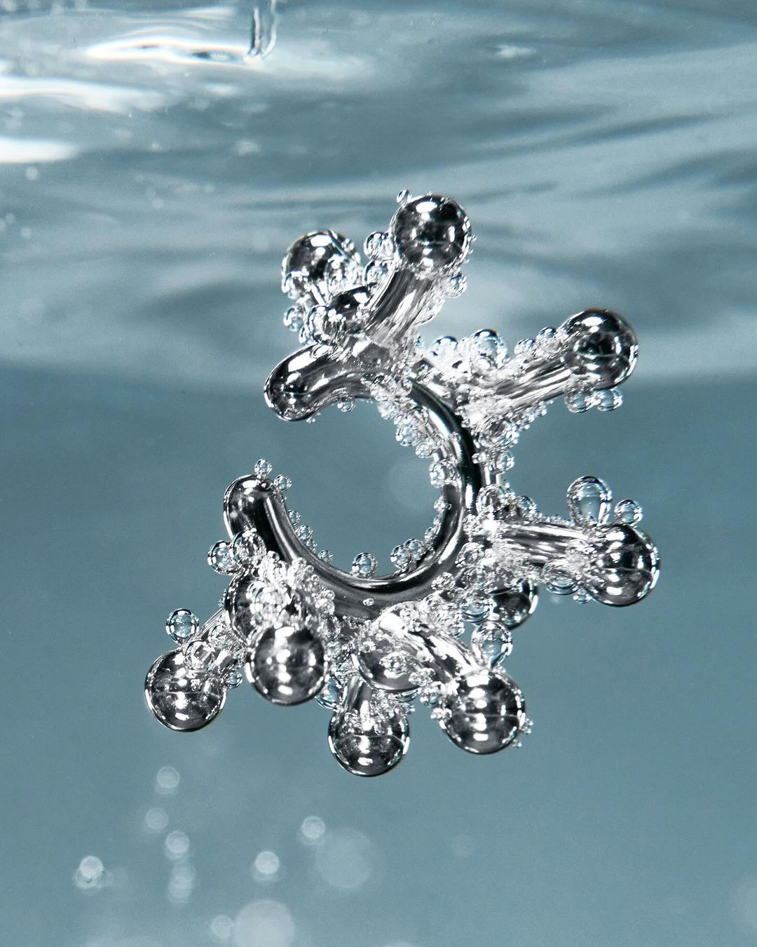 A silver metal jack toy is suspended in the center of light blue water filled with tiny clear bubbles in an eye-level shot.