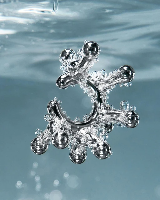 A silver metal jack toy is suspended in the center of light blue water filled with tiny clear bubbles in an eye-level shot.