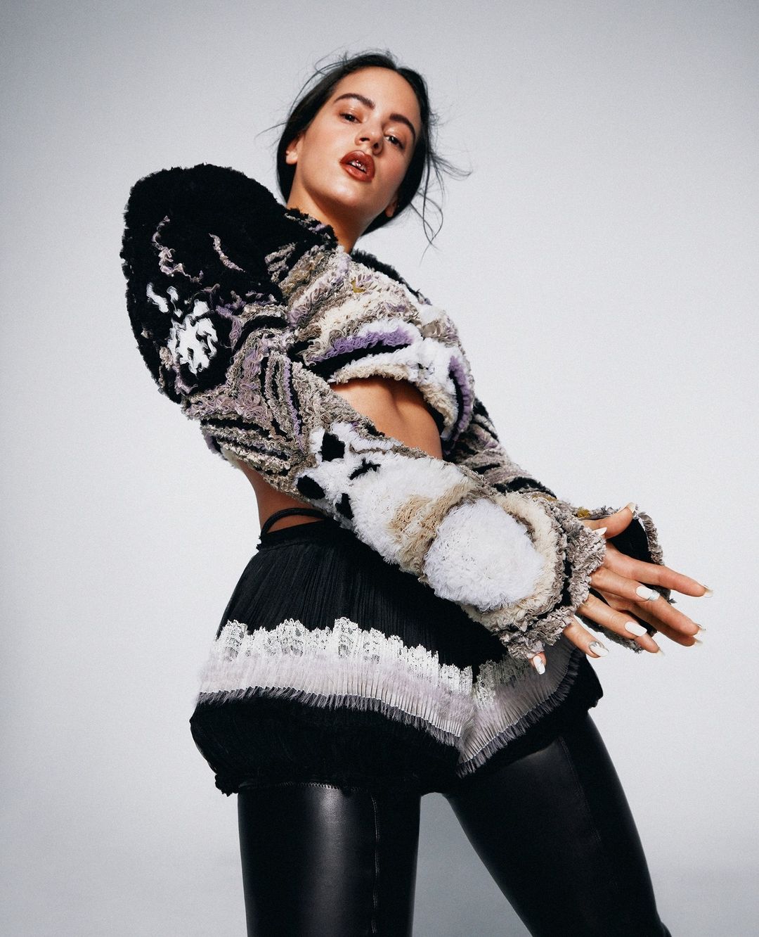 Woman with long dark hair poses in an embellished crop top and leggings in a fashion forward portrait against a neutral backdrop.
