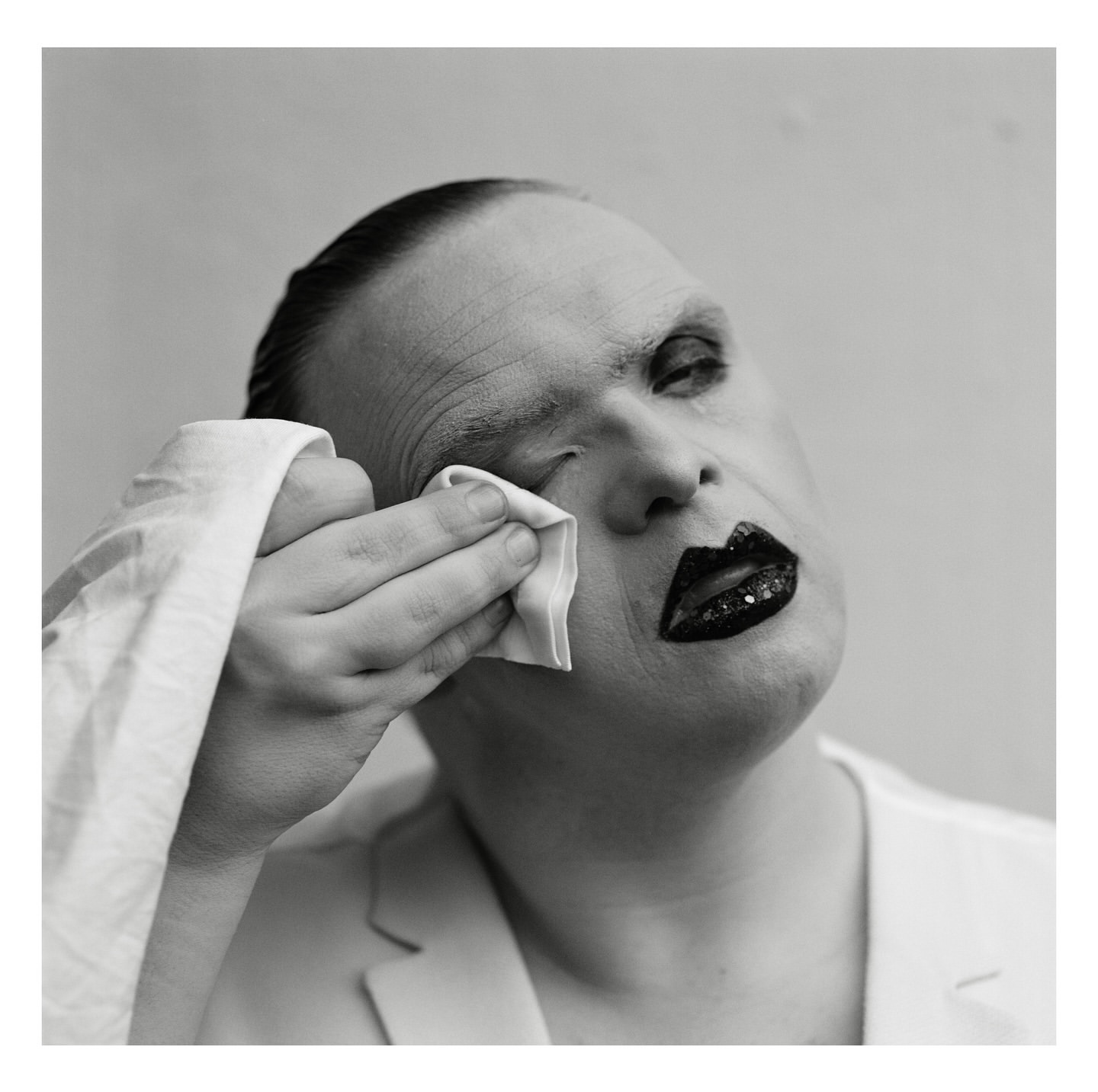 Close-up, black-and-white portrait of a person with heavy makeup wiping their eye with a white cloth, wearing a white jacket.